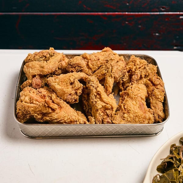 A metal tray overflowing with golden-brown fried chicken pieces, with a side of collard greens.