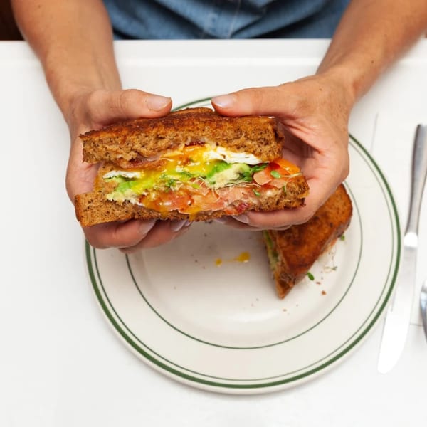 Hands holding a toasted sandwich with egg, bacon, avocado, and tomato over a plate.