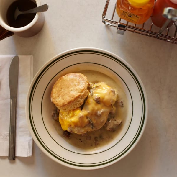 A bowl of biscuits and gravy with melted cheese, a coffee cup, and honey bottles.